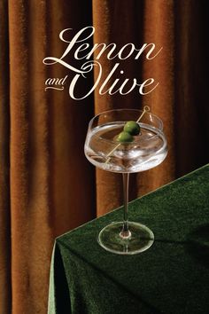 lemon and olive in a martini glass on a green tablecloth with curtains behind it