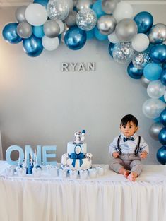 a baby boy sitting on top of a table next to a blue and white cake