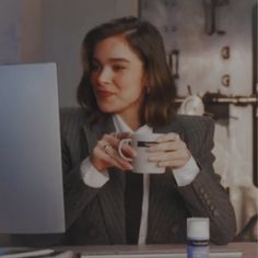 a woman sitting at a desk holding a coffee cup and looking at a computer screen