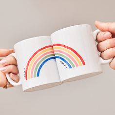 two hands holding coffee mugs with a rainbow on the front and side, one is white