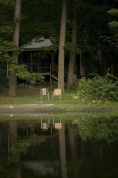 two lawn chairs sitting on the shore of a lake in front of a log cabin