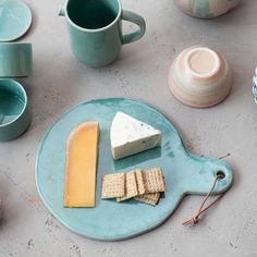 a blue plate with crackers and cheese on it next to cups, mugs and saucers