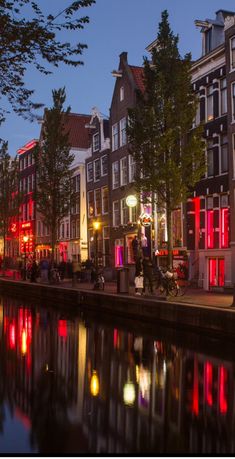 people are walking on the sidewalk near some buildings and water at night with red lights reflecting in the water