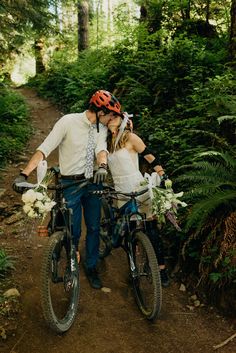 a man and woman kissing while riding bikes in the woods with greenery behind them