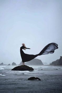 a woman standing on top of a rock in the ocean with her arms spread out