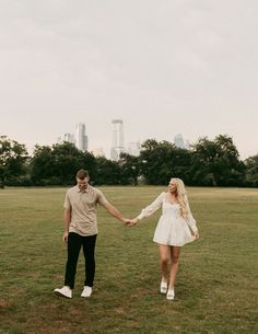 a man and woman holding hands while standing in a field with the city skyline behind them