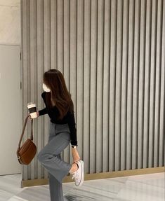 a woman in grey pants and black shirt holding a brown purse while standing next to a wall