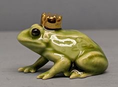 a green frog with a gold crown on its head sitting on a table in front of a gray background