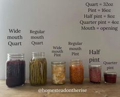 jars filled with different types of pickles sitting on top of a wooden table in front of a white wall