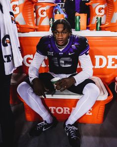 a football player sitting on a bench in front of orange coolers and holding a drink