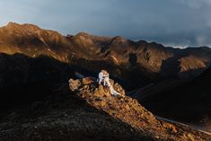 a bride and groom sitting on top of a mountain at sunset with mountains in the background