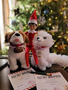 an elf and two stuffed dogs sitting next to each other on a desk in front of a christmas tree