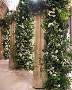 an arch covered in white flowers and greenery next to a stone wall with columns