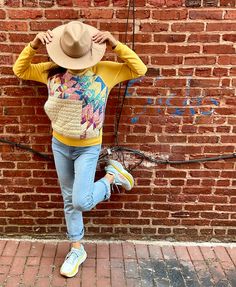 a woman standing in front of a brick wall with her hands on her head and wearing a hat