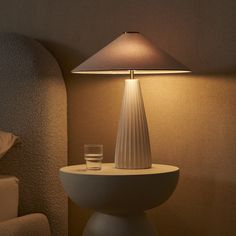 a white table lamp sitting on top of a night stand next to a glass filled with water