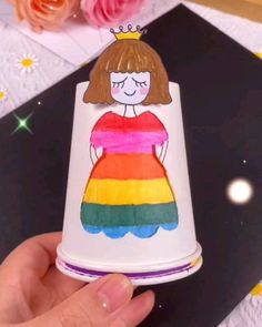 a hand holding a paper cup with a drawing of a girl wearing a rainbow dress