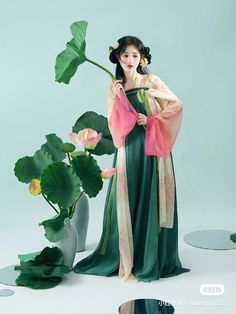 a woman in a long green dress holding a flower and standing next to a potted plant