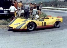 a yellow race car driving down a road with people standing around the side of it