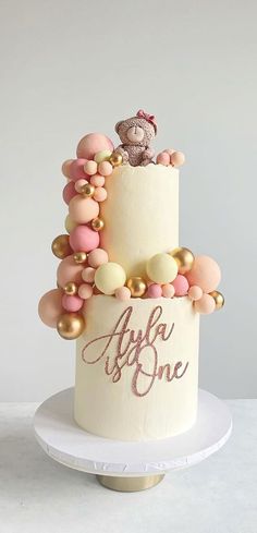 there is a white cake with pink and gold decorations on the top, topped with a teddy bear