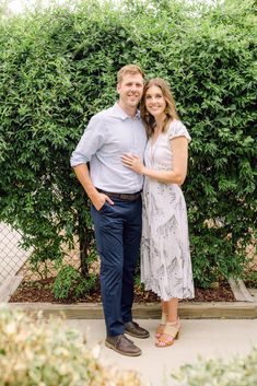 a man and woman standing next to each other in front of some bushes, smiling at the camera