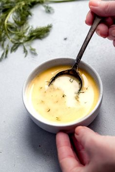 two hands holding a spoon over a bowl of soup with herbs on the table next to it