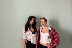 two young women standing next to each other in front of a gray wall and one is wearing a red leather jacket