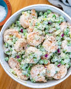 a bowl filled with shrimp and pea salad on top of a wooden table next to a spoon