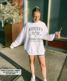 a woman is walking down the sidewalk with her tennis racket in her hand while wearing a white sweatshirt