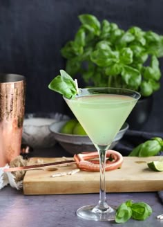 a green drink in a martini glass on a cutting board with basil leaves and copper cups