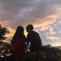 two people standing on top of a skateboard in front of a cloudy sky at sunset