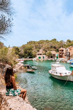 a woman sitting on the edge of a cliff looking at boats floating in the water