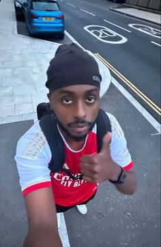 a man with a black hat and red shirt pointing at the camera while standing in front of a street