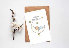 a greeting card with a baby sleeping on the moon next to some white flowers and a brown envelope