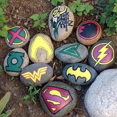 some rocks with different designs on them in the dirt near plants and flowers, one has a batman symbol painted on it