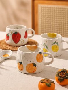 four oranges are sitting on a table next to two cups and spoons with fruit painted on them