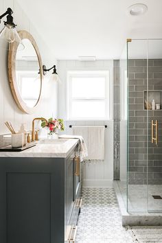 a bathroom with gray and white tile, marble counter tops, gold fixtures and a round mirror