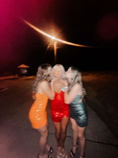 three women in short dresses hugging each other on the street at night with bright lights behind them