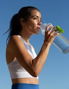 a woman drinking from a water bottle while standing in front of a blue cloudy sky