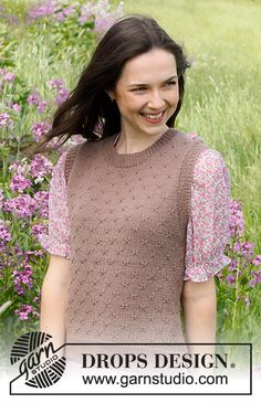 a woman standing in front of purple flowers wearing a brown knitted sweater and smiling at the camera