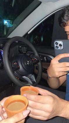 two people sitting in a car using their cell phones and drinking coffee from mugs