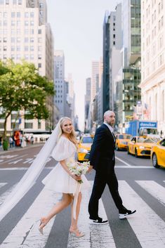 a bride and groom walking across the street in new york city, with taxi cabs behind them