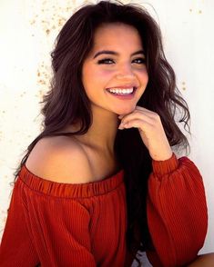 a woman with long hair smiling and wearing an off the shoulder red top in front of a white wall