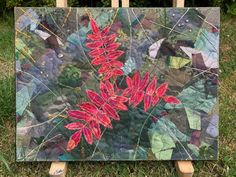 a painting with red flowers on it sitting in the grass next to a wooden easel