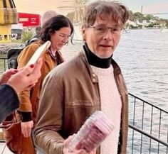 an older man standing next to a woman on a pier near the water and holding something in his hand