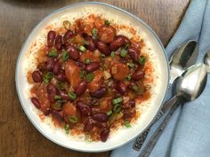 a white plate topped with beans and rice next to a silver spoon on top of a wooden table