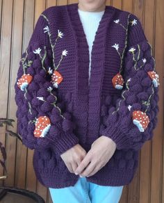 a woman standing in front of a wooden wall wearing a purple sweater with flowers on it