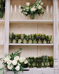 vases and glasses are arranged on an old dresser in front of a shelf filled with greenery