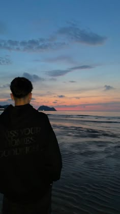 a man standing on top of a beach next to the ocean at sunset with clouds in the sky