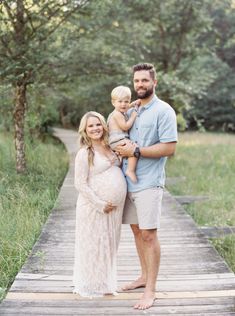 a pregnant couple and their son pose for a photo on a wooden walkway in the woods