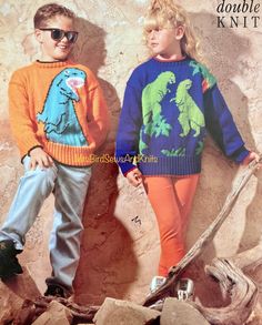 two children wearing sweaters and jeans standing next to each other in front of a rock wall